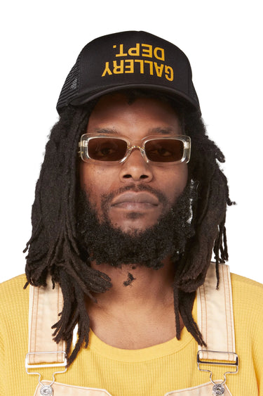 Model wearing a black trucker hat with a curved bill. The front displays an upside-down yellow "GALLERY DEPT." English logotype. The model is wearing tinted sunglasses, a yellow shirt, and light-colored overalls.