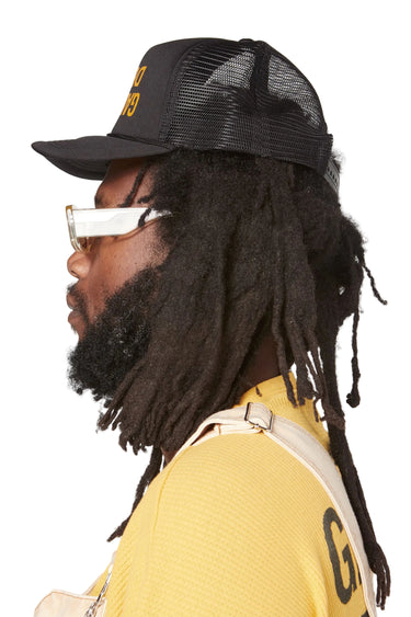 Right side profile of a model wearing the black mesh trucker hat. The curved bill extends forward, and the yellow front logo is partially visible. The mesh side panel and the model's long dreadlocks are visible.