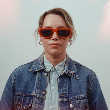 Front facing cropped image of female model in red sunglasses, denim jacket, denim shirt, and hoop earrings on a grey background.