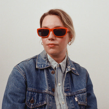 Side profile cropped image of female model in red sunglasses, denim jacket, denim shirt, and hoop earrings on a grey background.