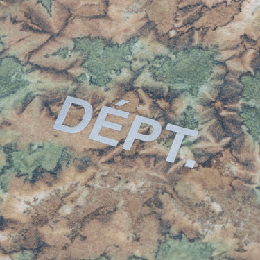 Close-up of the reflective silver “DÉPT.” logotype on the front of a camouflage-print athletic T-shirt, highlighting the lettering against the brown, olive green, and cream patterned fabric