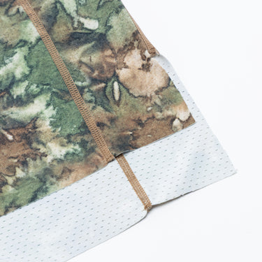 Close-up of the lower side hem of a camouflage-print athletic T-shirt, showing brown contrast stitching, the overlapping white perforated mesh panel, and the textured breathable fabric. Displayed against a plain white background.