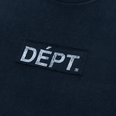 Close-up, angled detail shot of the front center of a black sleeveless tee against a dark fabric background, highlighting the rectangular black patch sewn onto the garment with distressed white “DÉPT.” lettering. The textured print and subtle faded wash are visible across the fabric and graphic.