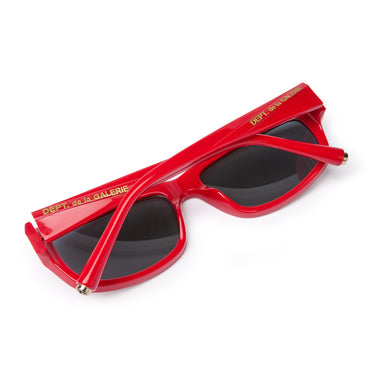 Red rectangular sunglasses with dark gray lenses folded face down, 'DEPT. de la GALERE' embedded logo in gold on the temple arm, silver 'G-Ball' studs on the ear tip, shown on a plain white background.