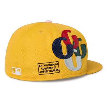 A three-quarter view of a yellow flat-brim cap is shown. The cap features multicolored G patch appliquéd lettering, a white New Era logo, and a black graphic that reads “ART ON DISPLAY COURTESY OF JOSUÉ THOMAS” near the back. The cap is photographed against a white background.
