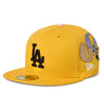 Three-quarter front view of a yellow flat-brim cap, featuring a black embroidered “LA” logo on the front, multicolored appliquéd "G" patch on the side, a white New Era logo, and a sticker on the brim, photographed against a white background.