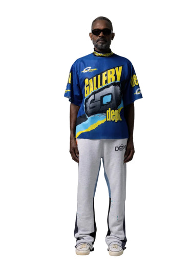 Full-body view of model wearing black sunglasses, a blue short sleeve mock neck jersey with oversized "GALLERY DEPT." graphics, light grey sweatpants, and black shoes.