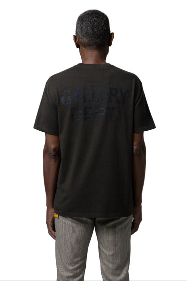 A man viewed from behind wearing a dark charcoal gray short-sleeve t-shirt with 'GALLERY DEPT.' printed in tonal dark lettering on the back, paired with light gray patterned trousers, against a white background.