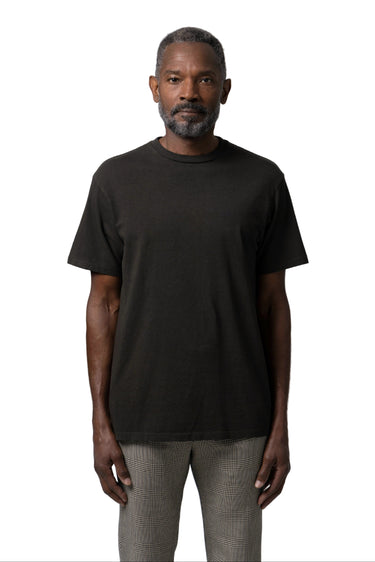 A man wearing a plain black short-sleeve crewneck t-shirt and light gray trousers, standing against a white background.