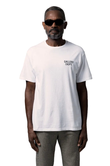 A man wearing a white short-sleeve t-shirt with 'GALLERY DEPT.' printed in black on the left chest, paired with gray trousers and black sunglasses, against a white background.