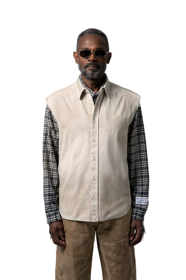 Male model wearing a light beige sleeveless overshirt over a black and white plaid button-down shirt, paired with brown pants and dark sunglasses, on a white background.