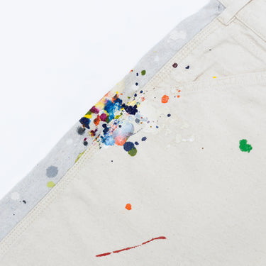 Angled close-up front view of the off-white shorts against a white background, highlighting multicolored paint splatters across the front pocket seam and contrasting light gray side panel, with visible contrast stitching and scattered