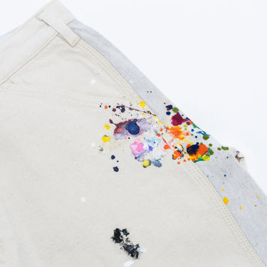 Angled close-up front view of the off-white shorts against a white background, showing multicolored paint splatters across the back pocket and contrasting light gray side panel, with visible stitching and scattered paint marks.