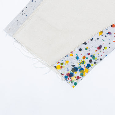Angled close-up of the shorts against a white background, highlighting the frayed off-white hem, exposed threads, and contrasting light gray fabric panel covered in multicolored paint splatters