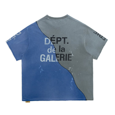 Back view of a two-tone T-shirt laid flat against a white background, featuring blue fabric on the left and charcoal gray on the right, separated by curved black stitching. An enlarged “DÉPT. de la GALERIE” logotype spans the back in contrasting black and white lettering, with faded paint marks and partial sleeve graphics.