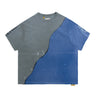 Front view of a short-sleeve T-shirt laid flat against a white background, featuring a two-tone design with a faded charcoal-gray upper and left section and a blue right and lower section, curved diagonal black stitching across the center, scattered paint marks, and partial graphic lettering on both sleeves.
