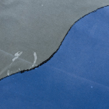Angled close-up of the T-shirt, showing the curved black overlock stitching joining the charcoal-gray and blue fabric panels, with scattered light-colored paint marks and the garment’s washed texture.
