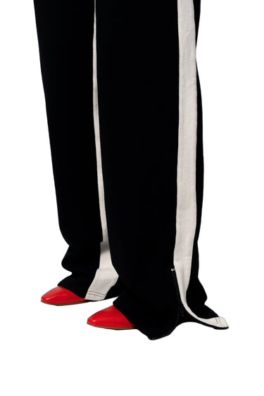 Black wide-leg trousers with a bold white vertical stripe down the outer side of each leg, zipper detail on bottom, worn with bright red pointed-toe shoes on a white background.
