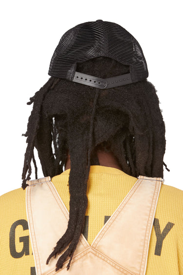 Rear view of a model wearing the black trucker hat, showing the black mesh back panels and adjustable snap closure. The model's long dreadlocks extend below the hat.